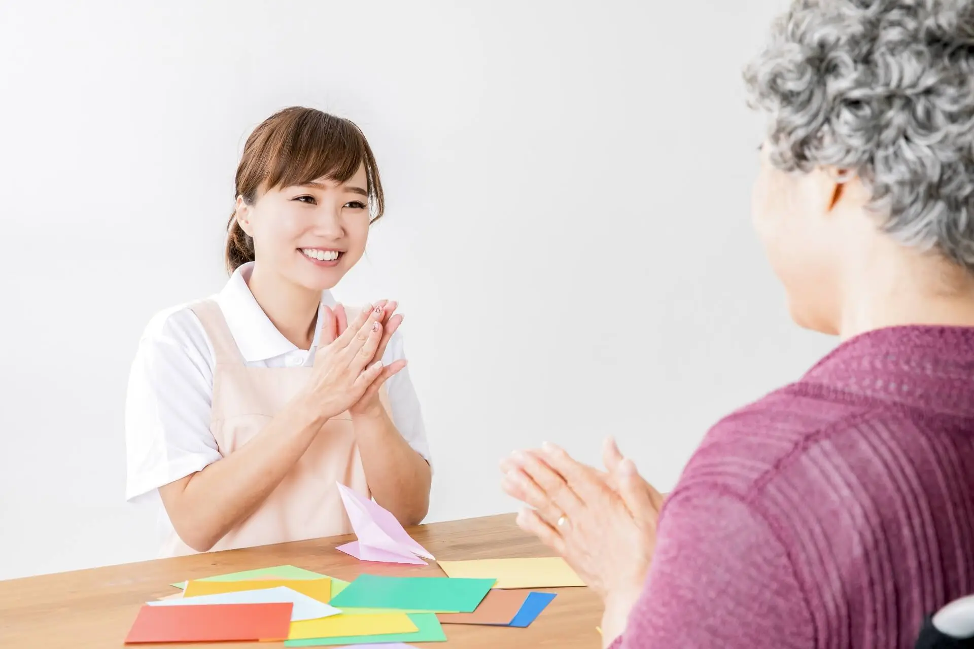 車椅子に乗ったご高齢の女性と、女性介護スタッフが机に座り、共に折り紙を楽しんでいる様子の写真